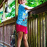 child, girl, smiling, outdoor, porch, wooden_railing, greenery, summer_clothing, shorts, tshirt, happy, nature, daylight, sandals, balcony, playful, cute, sunlight, wooden_floor, casual