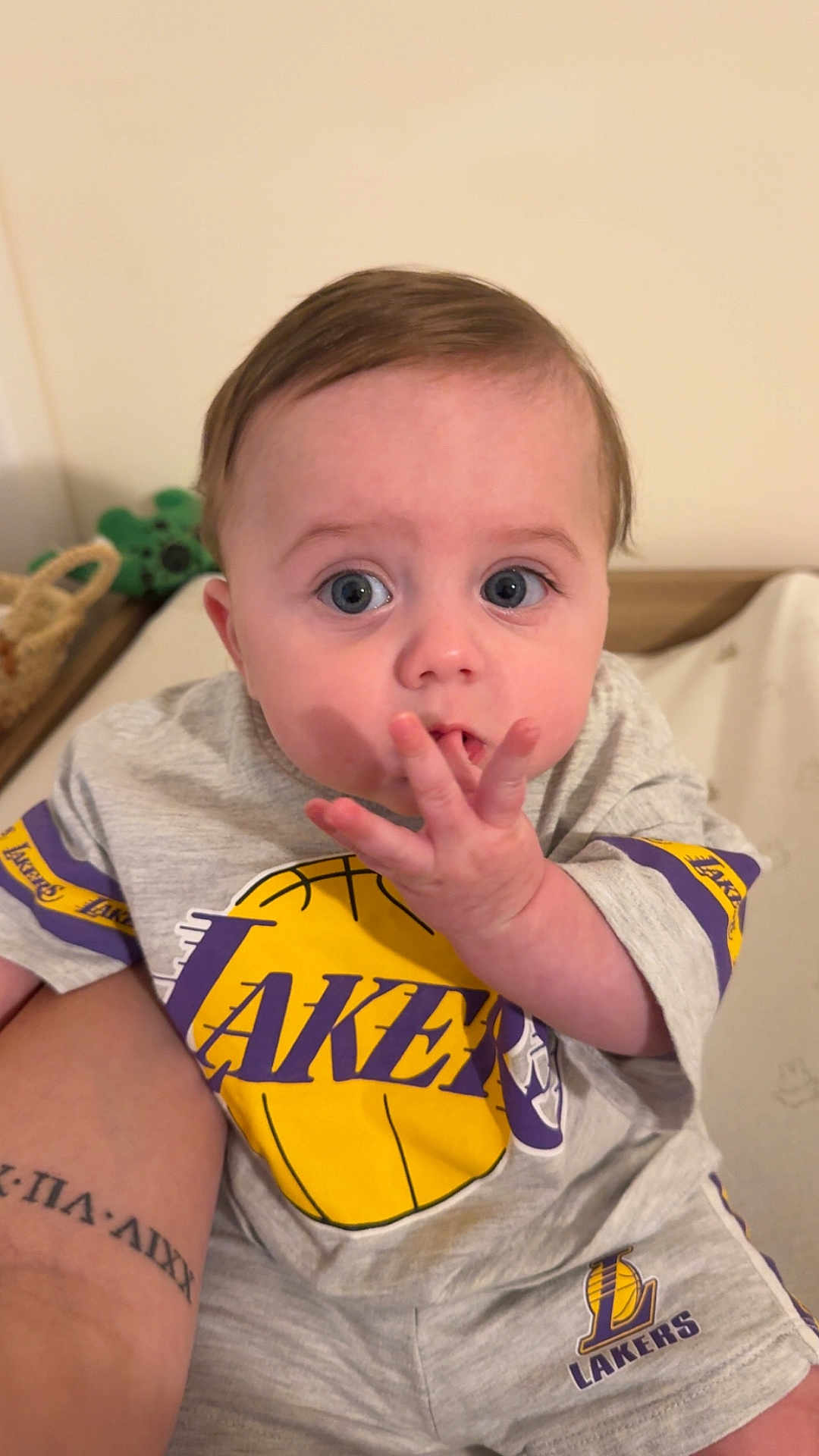 Pablo participe au concours pour gagner de l'argent avec cette photo : baby, child, lakers, clothing, grey_outfit, basketball_logo, indoor, person, hand, face, blue_eyes, short_hair, tattoo, sitting, cute, infant, looking_up, casual, home, portrait