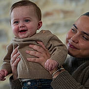 Pablo a rejoint le concours — aidez-le/la à gagner de superbes lots ! adult, baby, beige_sweater, blue_jeans, casual_wear, closeup, cozy_clothing, face, family, hands, happy, holding, indoor, joy, love, person, portrait, smile, stone_wall, warm_lighting