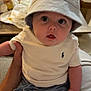 baby, child, hat, bucket_hat, white_shirt, blue_shorts, indoor, hand, curious, face, person, clothing, casual, seated, infant, cute, portrait, expression, human, young