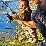 child, toddler, man, fishing_rod, fishing, water, lake, outdoor, nature, grass, tree_branches, adult, bonding, parent_child, holding, curious, casual_clothing, daylight, recreation, hobby