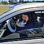 woman, cat, kitten, car, inside_car, smiling, holding, seatbelt, steering_wheel, daylight, outdoor, parking_lot, trees, blue_sky, casual_clothing, happy, pet, animal, human, window