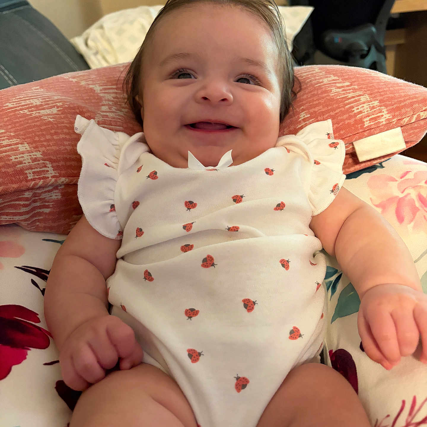 Sofia is registered to the contest to win money with this photo: baby, infant, smile, onesie, ladybug_pattern, cushion, pillow, indoor, happy, cute, child, person, furniture, floral, skin, head, arm, leg, cheerful, comfort