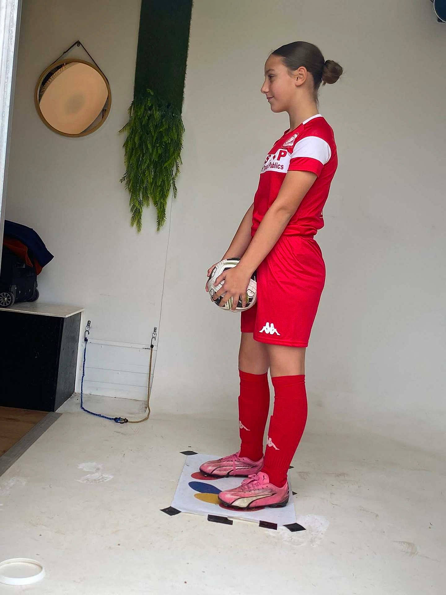 athlete, casual, child, floor, hair_bun, indoor, mirror, plant, pose, ready, red_clothing, red_socks, side_view, soccer, soccer_ball, sports_shoes, sports_uniform, standing, wall, young_girl