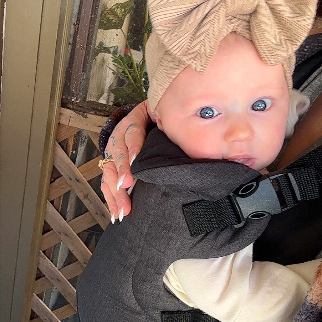 Beverly is registered to the contest to win money with this photo: baby, baby_carrier, blue_eyes, bow, clothing, cute, fabric, face, fingers, hand, headband, holding, indoor, infant, lattice, person, plant, skin, straps, tattoo