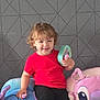 black_pants, cartoon_character, child, colorful, curly_hair, cute, furniture, geometric_wall, happy, home, indoor, person, playful, plush_chair, red_shirt, sitting, smile, toddler, toy, young_child