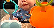 Bryson joined the competition — help win amazing prizes! baby, infant, toy, colorful, face, curious, expression, blue_clothing, elephant_toy, orange_toy, green_ring, blue_ring, indoor, carpet, basket, playtime, child, patterned_cushion, closeup, cute