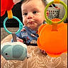 baby, infant, toy, colorful, face, curious, expression, blue_clothing, elephant_toy, orange_toy, green_ring, blue_ring, indoor, carpet, basket, playtime, child, patterned_cushion, closeup, cute