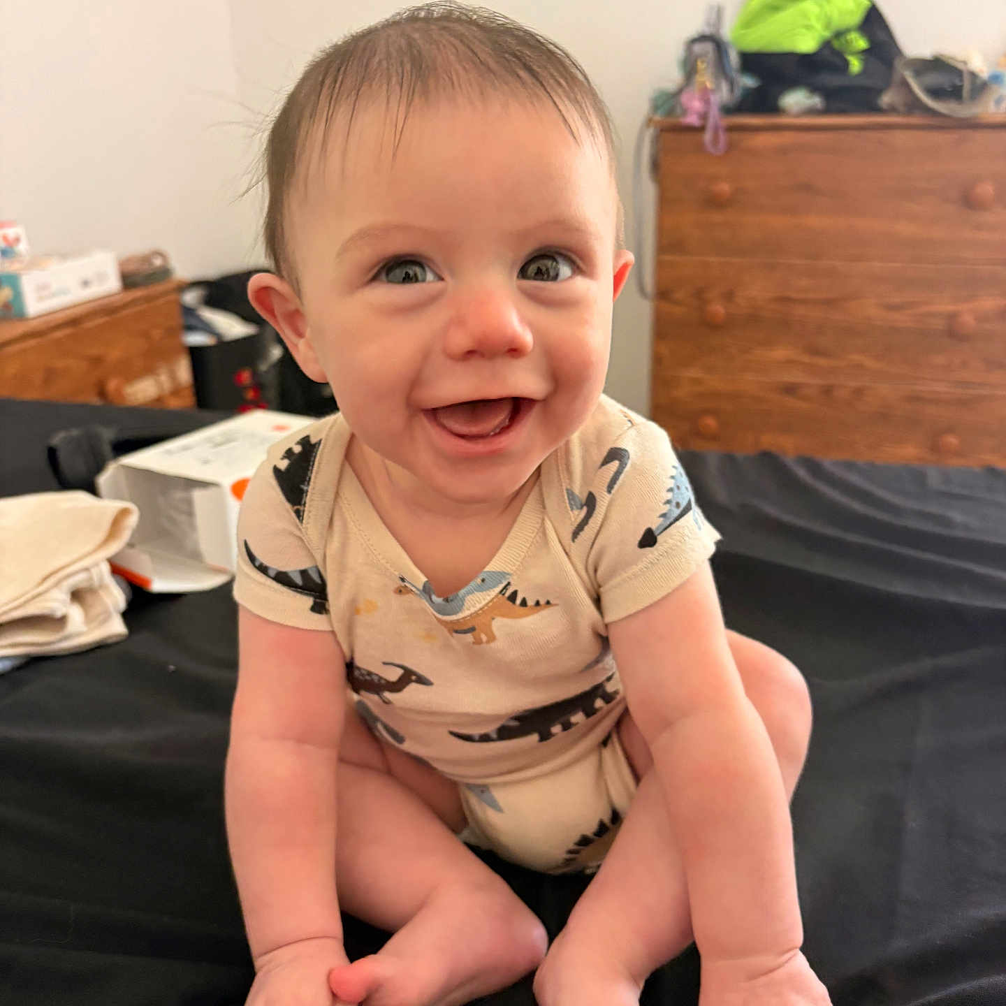 Bryson is registered to the contest to win money with this photo: baby, smiling, dinosaur_onesie, infant, sitting, bed, black_bedspread, wooden_dresser, clothing, indoors, cute, happy, child, person, face, skin, hair, room, furniture, casual