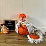 baby, pumpkin, orange_hat, blanket, doll, sign, wooden_floor, white_wall, halloween, cute, infant, costume, holiday, indoors, child, smiling, decor, festive, portrait, seasonal