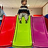 Naël participe au concours pour gagner de l'argent avec cette photo : child, slide, indoor_play_area, colorful, red_slide, green_slide, purple_slide, toddler, barefoot, striped_sweater, gray_pants, ceiling_tiles, soft_flooring, playground, young_child, curious, waiting, fun, recreation, leisure