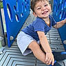 child, toddler, smiling, playground, blue, shirt, shorts, footwear, sitting, happy, outdoor, metal_floor, play_structure, young_child, casual_clothing, cute, fun, daylight, person, joy
