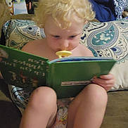 Madilyn is registered to the contest to win money with this photo: toddler, child, book, reading, pacifier, couch, cushion, blond_hair, indoor, patterned, diaper, curly_hair, seated, furniture, home, young_child, concentration, comfort, blanket, pillow