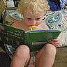 toddler, child, book, reading, pacifier, couch, cushion, blond_hair, indoor, patterned, diaper, curly_hair, seated, furniture, home, young_child, concentration, comfort, blanket, pillow