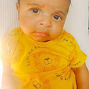 Isaiah is registered to the contest to win money with this photo: baby, child, infant, yellow_clothing, lion_print, pouty_expression, big_eyes, close_up, cushion, soft_lighting, indoors, cute, person, portrait, sitting, hand, curly_hair, bright_colors, toddler, young_child