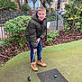 child, boy, smiling, outdoor, mini_golf, golf_club, green_jacket, boots, green_ball, grass, plants, trees, urban, fence, pavement, casual_clothing, daytime, recreation, sport, happy