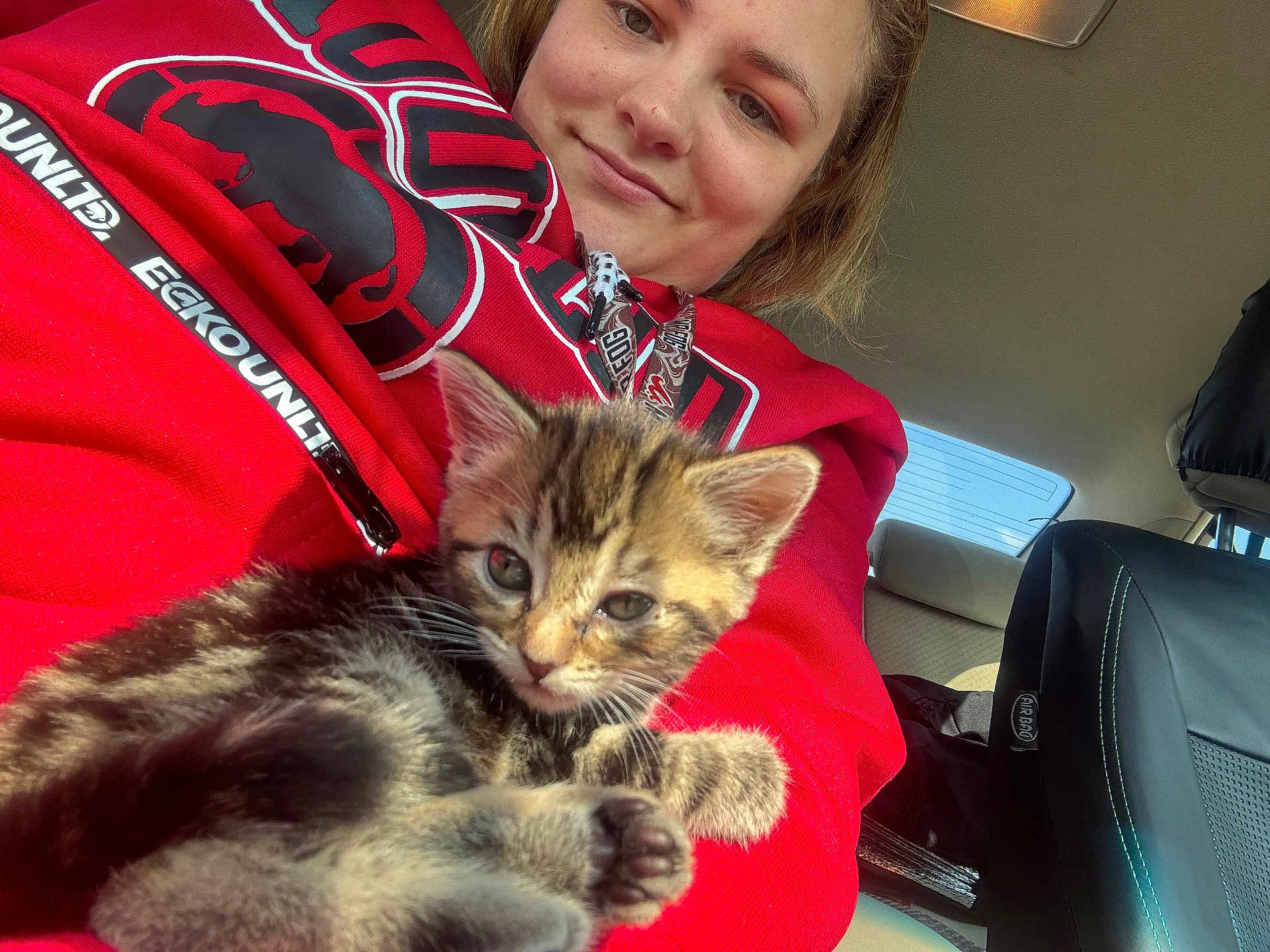 Stormy Mae is registered to the contest to win money with this photo: person, kitten, cat, animal, feline, indoor, car_interior, red_hoodie, smile, young_adult, closeup, pet, cute, cozy, seated, window, seat, seatbelt, daylight, casual