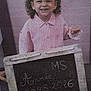 child, smiling, chalkboard, curly_hair, indoor, striped_shirt, handwritten_text, young, person, portrait, happy, clothing, sign, school, education, girl, standing, writing, background, holding