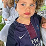 child, curly_hair, blue_headband, sports_jersey, paris_saint_germain, nike_logo, outdoor, grass, adult_in_background, pout, portrait, person, casual_clothing, earrings, greenery, daylight, expression, standing, close_up, fashion