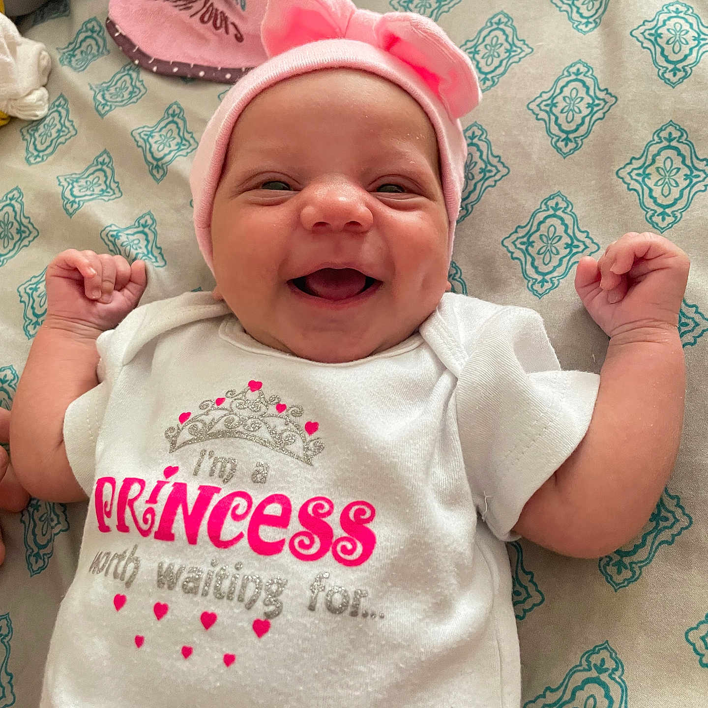 Bianca is registered to the contest to win money with this photo: baby, infant, smiling, pink_bow, onesie, text_on_clothing, bedspread, patterned, happy, cute, lying_down, indoor, child, person, headwear, small_hands, cheeks, portrait, closeup, clothing