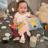 baby, child, smiling, toy, teddy_bear, bottle, fabric_book, cushion, cloud_pattern, indoor, happy, sitting, infant, playtime, soft_toys, footwear, white_shirt, purple_pants, baby_seat, home