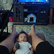 Victor is registered to the contest to win money with this photo: baby, infant, person, legs, feet, mittens, onesie, living_room, television, toy_house, cabinet, fan, floor, carpet, furniture, home_interior, relaxation, indoor, footwear_absent, screen