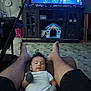 baby, infant, person, legs, feet, mittens, onesie, living_room, television, toy_house, cabinet, fan, floor, carpet, furniture, home_interior, relaxation, indoor, footwear_absent, screen