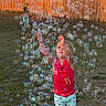 child, toddler, bubbles, outdoor, grass, fence, sunlight, playing, reaching, red_tank_top, shorts, ladybug_print, happy, summer, fun, joy, nature, colorful, daylight, activity