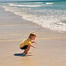 child, beach, sand, ocean, water, waves, yellow_shirt, shorts, barefoot, sunny, nature, outdoor, curious, squatting, shore, reflection, sky, sea, summer, vacation