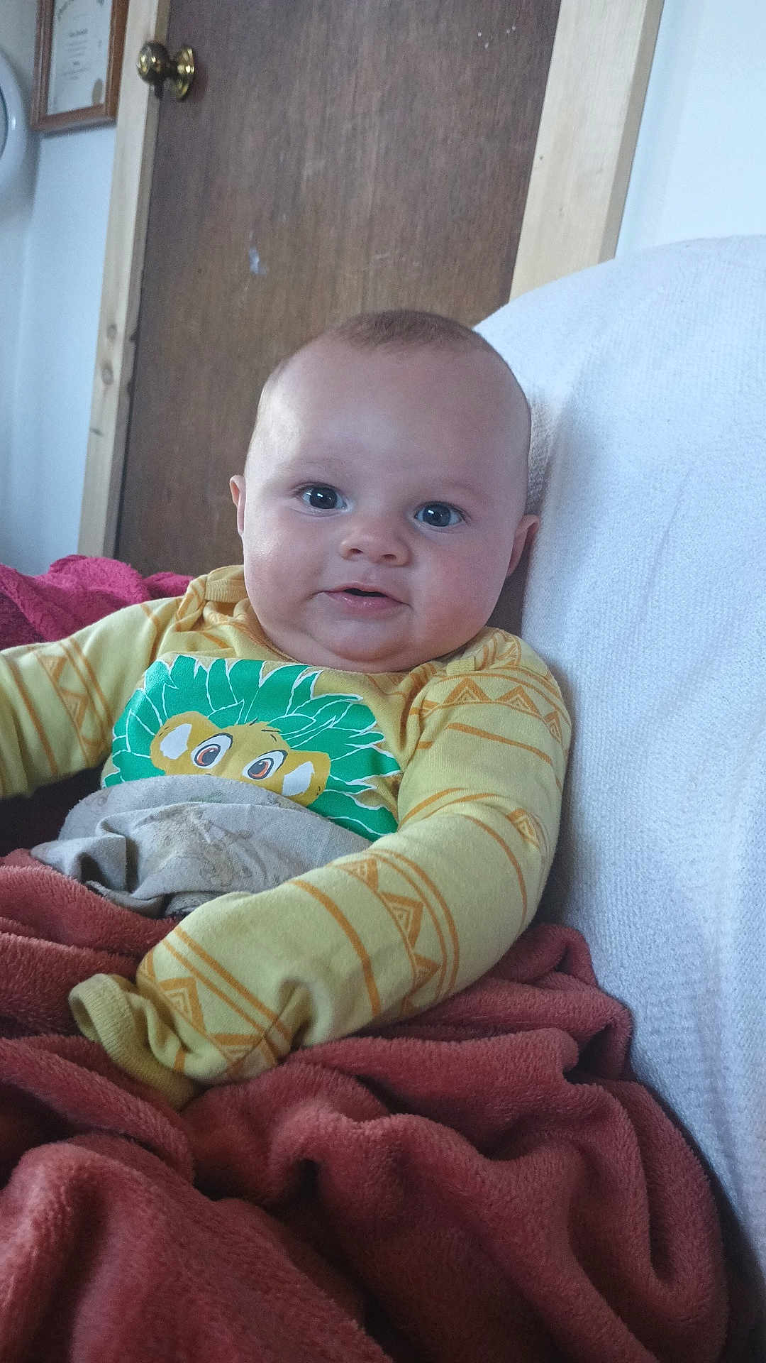 Shawn joined the competition — help win amazing prizes! baby, child, infant, face, smile, yellow_shirt, lion_graphic, blanket, red_blanket, couch, white_couch, indoors, door, wooden_door, person, cute, cozy, happy, portrait, seated