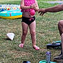 Violet is registered to the contest to win money with this photo: toddler, child, playing, water_balloon, grass, summer, outdoor, inflatable_pool, shoes, sandals, person, hand, park, tree, building, people, casual_clothing, fun, daytime, recreation