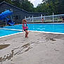 Violet is registered to the contest to win money with this photo: child, toddler, pool, life_jacket, water_slide, outdoor, play, concrete, water, summer, fence, trees, cloudy_sky, building, adult, recreation, fun, curly_hair, splash, swimwear