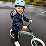 balance_bike, bike, child, green_bike, handlebar, hands, helmet, helmeted_child, jacket, outdoor, parking_lot, pavement, playful, portrait, rainy_day, smile, sneakers, toddler, wet_ground, wheel