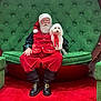 santa_claus, dog, white_dog, red_ribbon, green_chair, plush_furniture, red_carpet, holiday, festive, beard, glasses, boots, costume, seated, smiling, indoors, portrait, celebration, christmas, pet