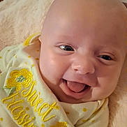 Nova is registered to the contest to win money with this photo: baby, infant, smiling, tongue_out, bib, embroidery, blanket, closeup, face, head, eyes, chubby_cheeks, happy, indoors, clothing, lying_down, newborn, skin, adorable, portrait