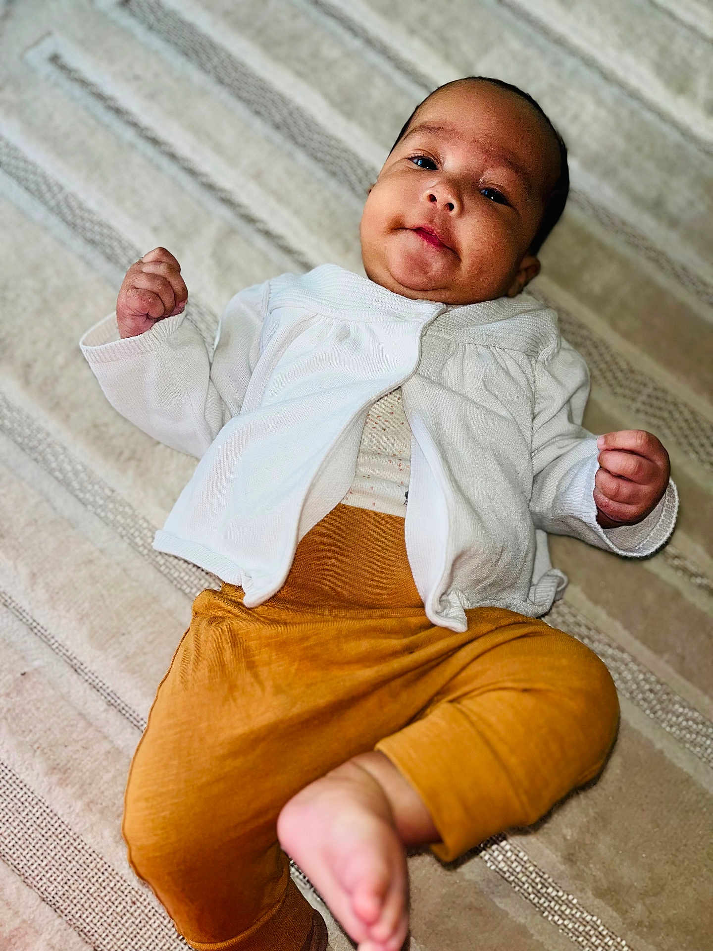 Fatoumata participe au concours pour gagner de l'argent avec cette photo : baby, infant, child, white_cardigan, mustard_pants, barefoot, lying_down, rug, patterned_rug, curious_expression, fists, indoors, portrait, cute, small_hands, smiling, looking_at_camera, cozy, soft_lighting, young_child