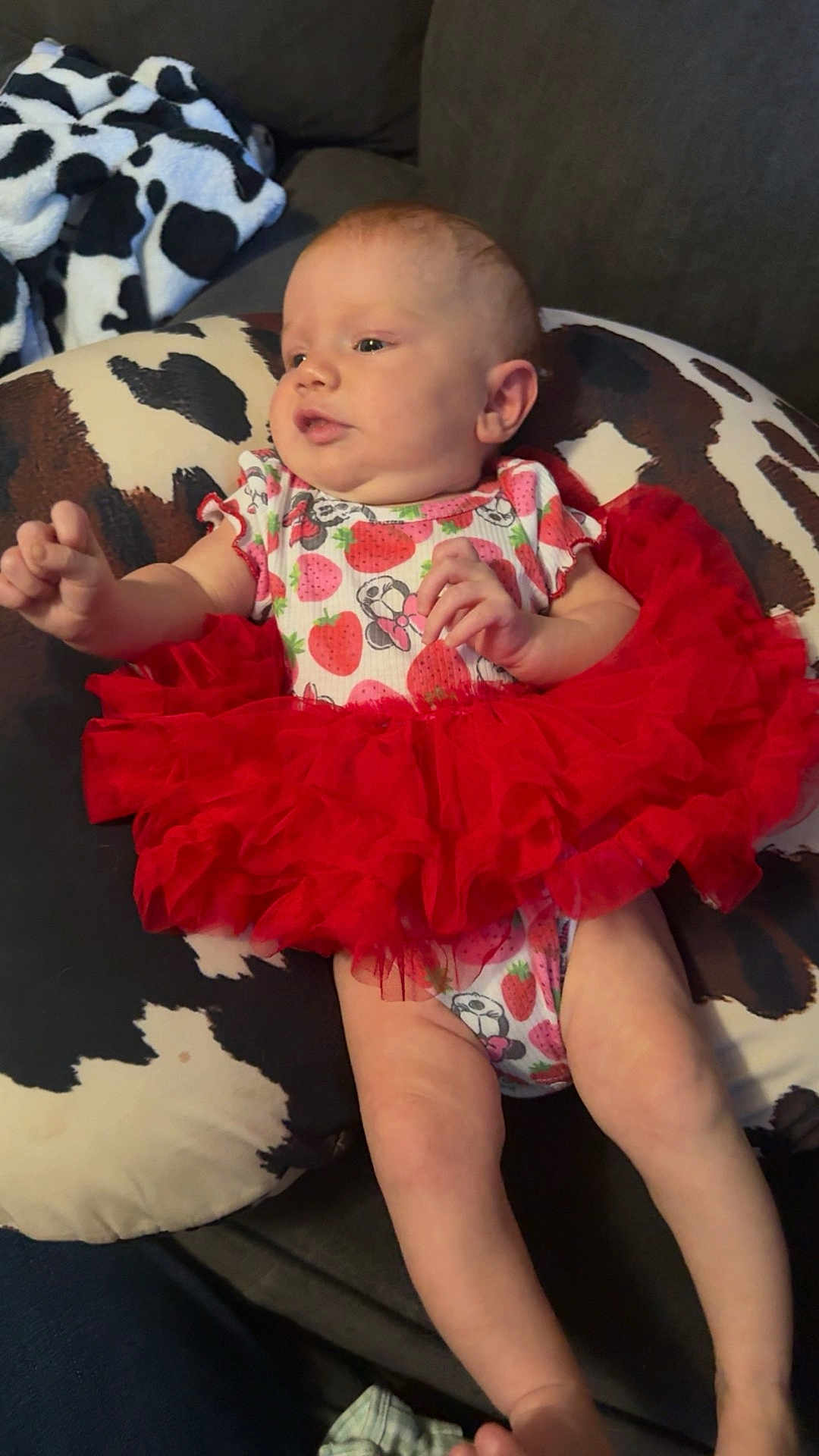 Oakley is registered to the contest to win money with this photo: baby, infant, tutu, red_skirt, onesie, strawberry_pattern, minnie_mouse, pillow, cow_print, couch, furniture, child, person, skin, legs, arms, head, face, indoor, relaxing