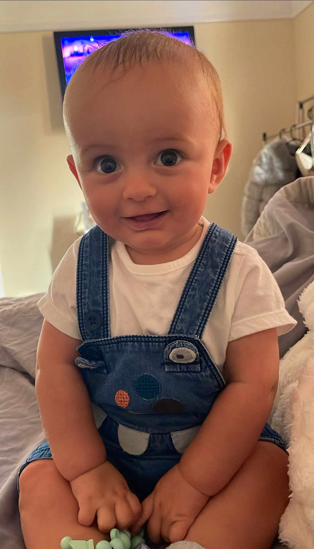 Arlo James is registered to the contest to win money with this photo: baby, blanket, casual_clothing, child, cozy, cute, denim_overall, face, hands, happy, indoors, infant, legs, portrait, room, sitting, smiling, soft_toy, toy, white_tshirt