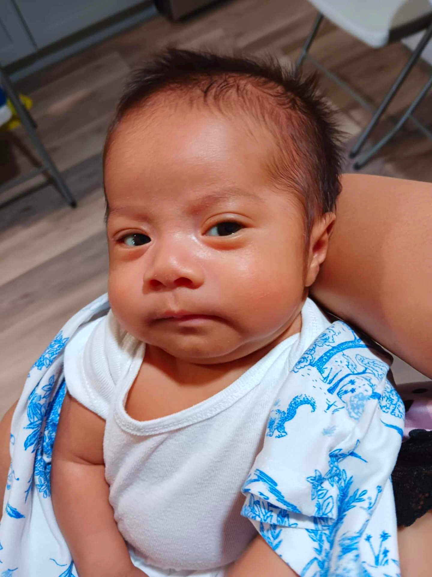 Sergio is registered to the contest to win money with this photo: baby, infant, child, face, eyes, arm, clothing, white_shirt, blue_pattern, blanket, indoor, floor, wooden_floor, person, portrait, newborn, cute, skin, expression, holding