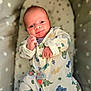 Tommy is registered to the contest to win money with this photo: baby, infant, onesie, dinosaur_pattern, smile, face, hand, cushion, star_pattern, sleeping_bed, cute, newborn, comfort, soft_toys, indoor, portrait, child, cozy, apparel, resting