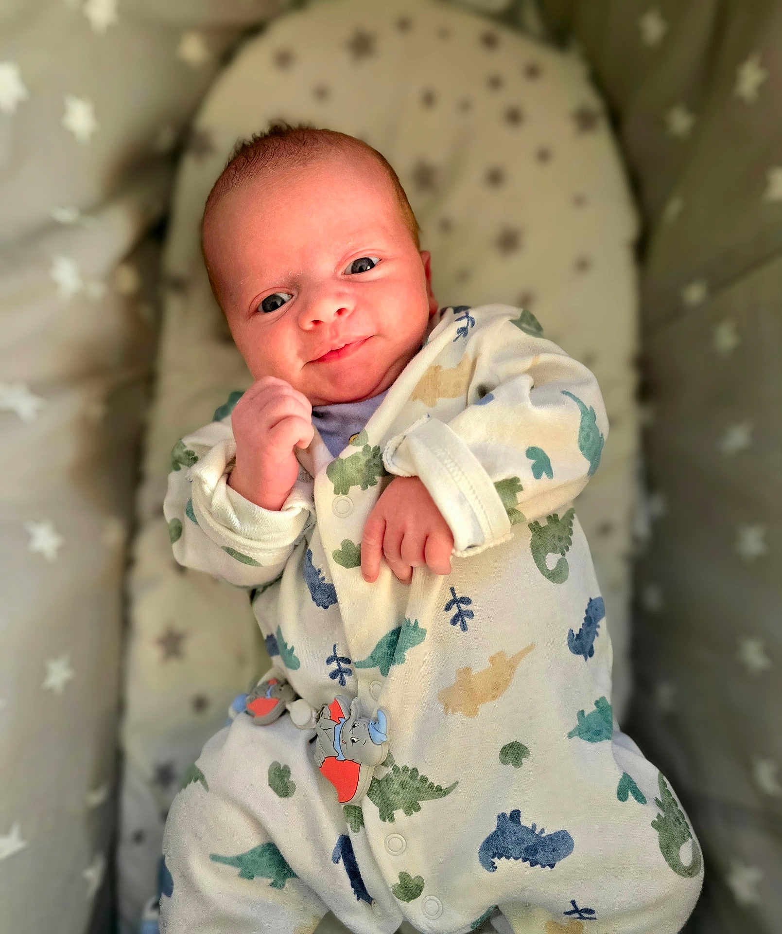 Tommy is registered to the contest to win money with this photo: baby, infant, onesie, dinosaur_pattern, smile, face, hand, cushion, star_pattern, sleeping_bed, cute, newborn, comfort, soft_toys, indoor, portrait, child, cozy, apparel, resting