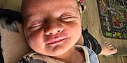 Tommy is registered to the contest to win money with this photo: baby, child, smile, sleeping, onesie, dinosaur_pattern, couch, beige, floor, playmat, wooden_floor, indoor, person, hand, foot, colorful_clothing, home, relaxation, portrait, infant