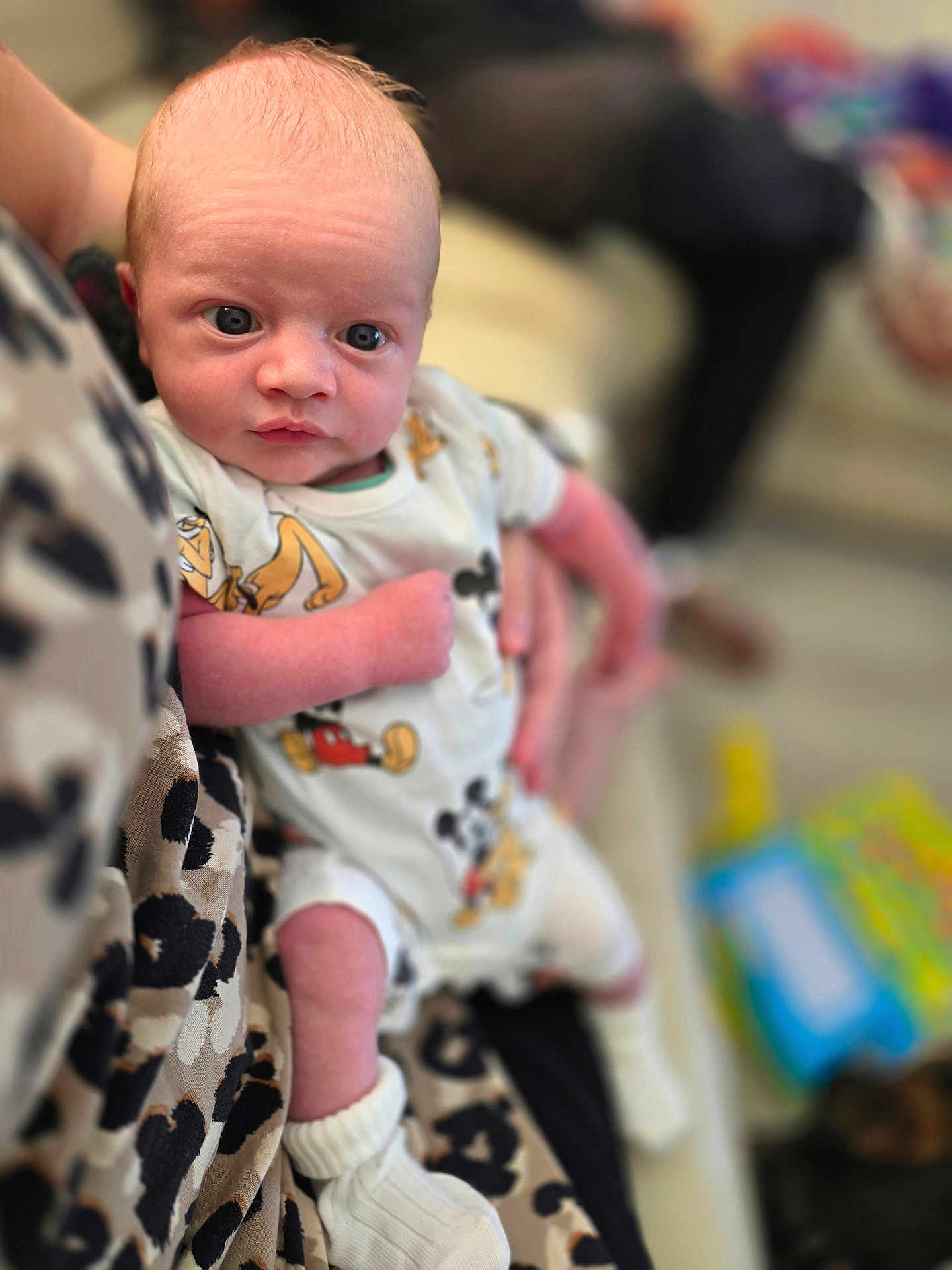 Tommy is registered to the contest to win money with this photo: baby, infant, person, child, mickey_mouse, onesie, clothing, socks, hand, holding, face, eyes, skin, blurred_background, couch, patterned_clothing, indoor, soft_light, cute, portrait