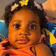 Janylah is registered to the contest to win money with this photo: baby, child, curly_hair, butterfly_clips, pink_clothing, cushion, patterned_cushion, indoor, portrait, closeup, face, eyes, warm_lighting, relaxed, cute, infant, person, young_child, headwear, smile
