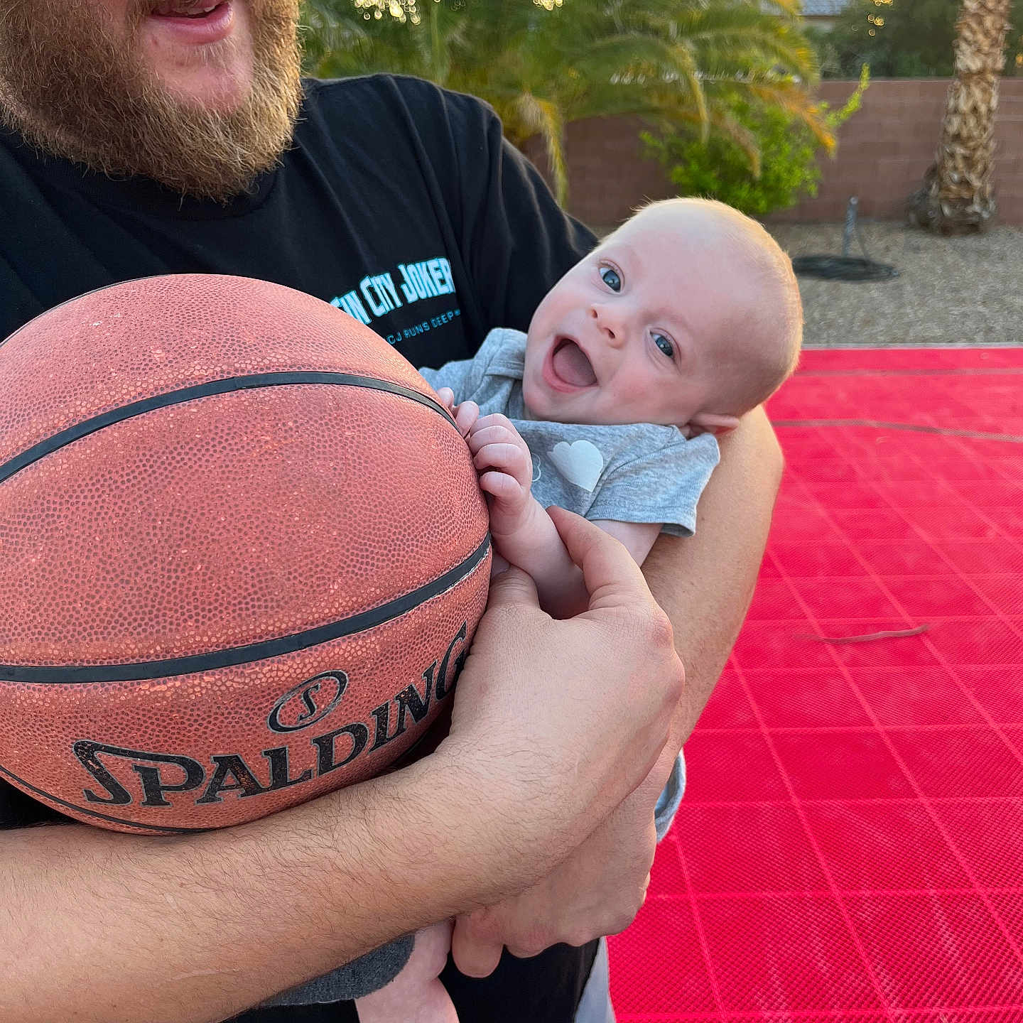 Collin is registered to the contest to win money with this photo: baby, ball, basketball, beard, court, grass, happy, holding, man, outdoor, palm_tree, person, playful, shirt, shorts, smile, sock, sport, sunset, tree