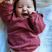 Céleste participe au concours pour gagner de l'argent avec cette photo : baby, infant, smiling, mouth_open, dark_hair, hair_clip, blanket, towel, pink_shirt, long_sleeves, beige_pants, lying_down, portrait, closeup, happy, chubby_cheeks, hand_raised, cute, indoor, floral_bedding