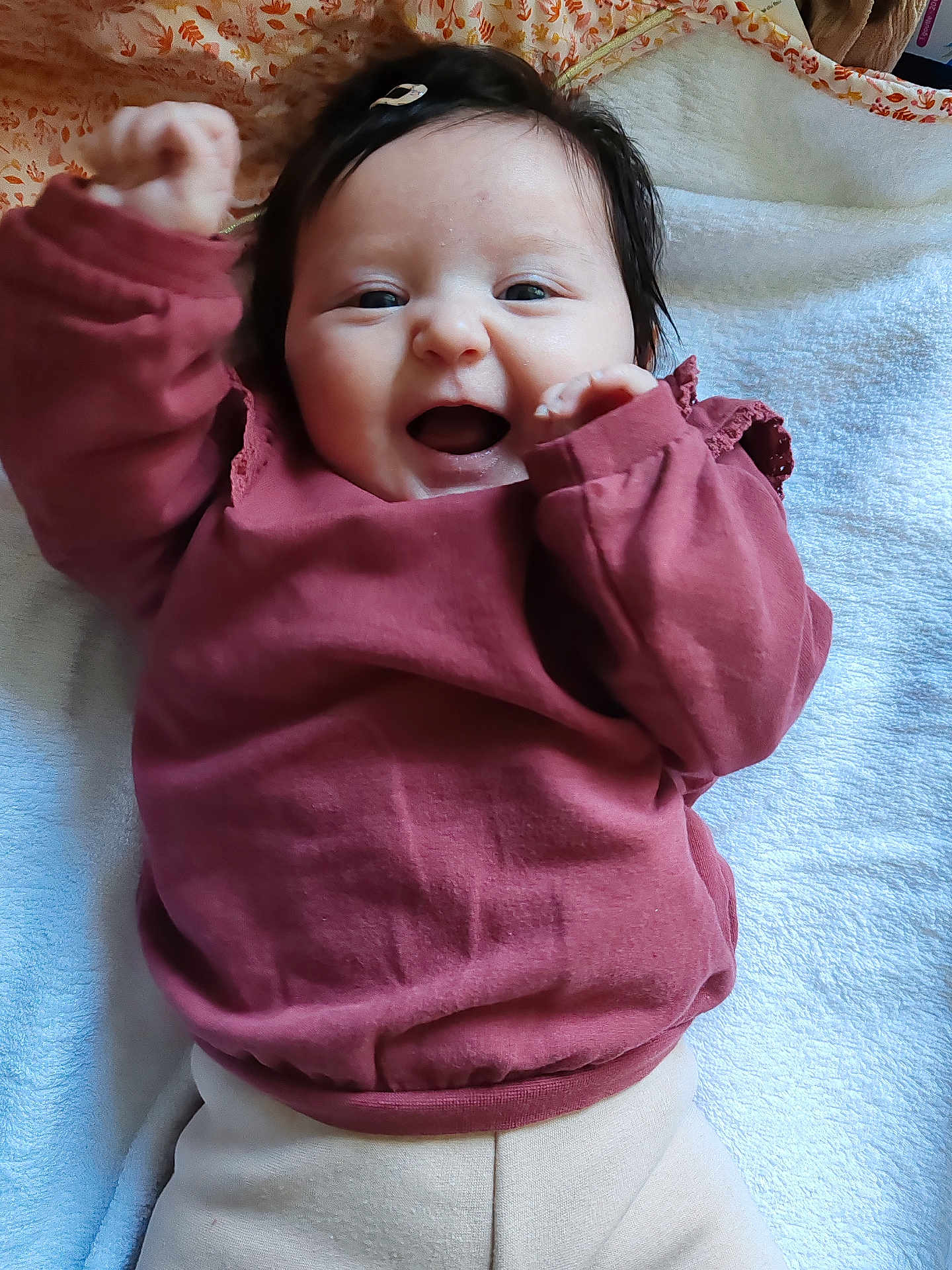 Céleste participe au concours pour gagner de l'argent avec cette photo : baby, infant, smiling, mouth_open, dark_hair, hair_clip, blanket, towel, pink_shirt, long_sleeves, beige_pants, lying_down, portrait, closeup, happy, chubby_cheeks, hand_raised, cute, indoor, floral_bedding