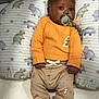 Karter is registered to the contest to win money with this photo: baby, infant, pacifier, orange_sweater, beige_pants, halloween_prints, dinosaur_pillow, cute, indoor, lying_down, child, soft_lighting, closeup, portrait, cozy, baby_clothes, baby_boy, comfort, sleeping_area, playpen