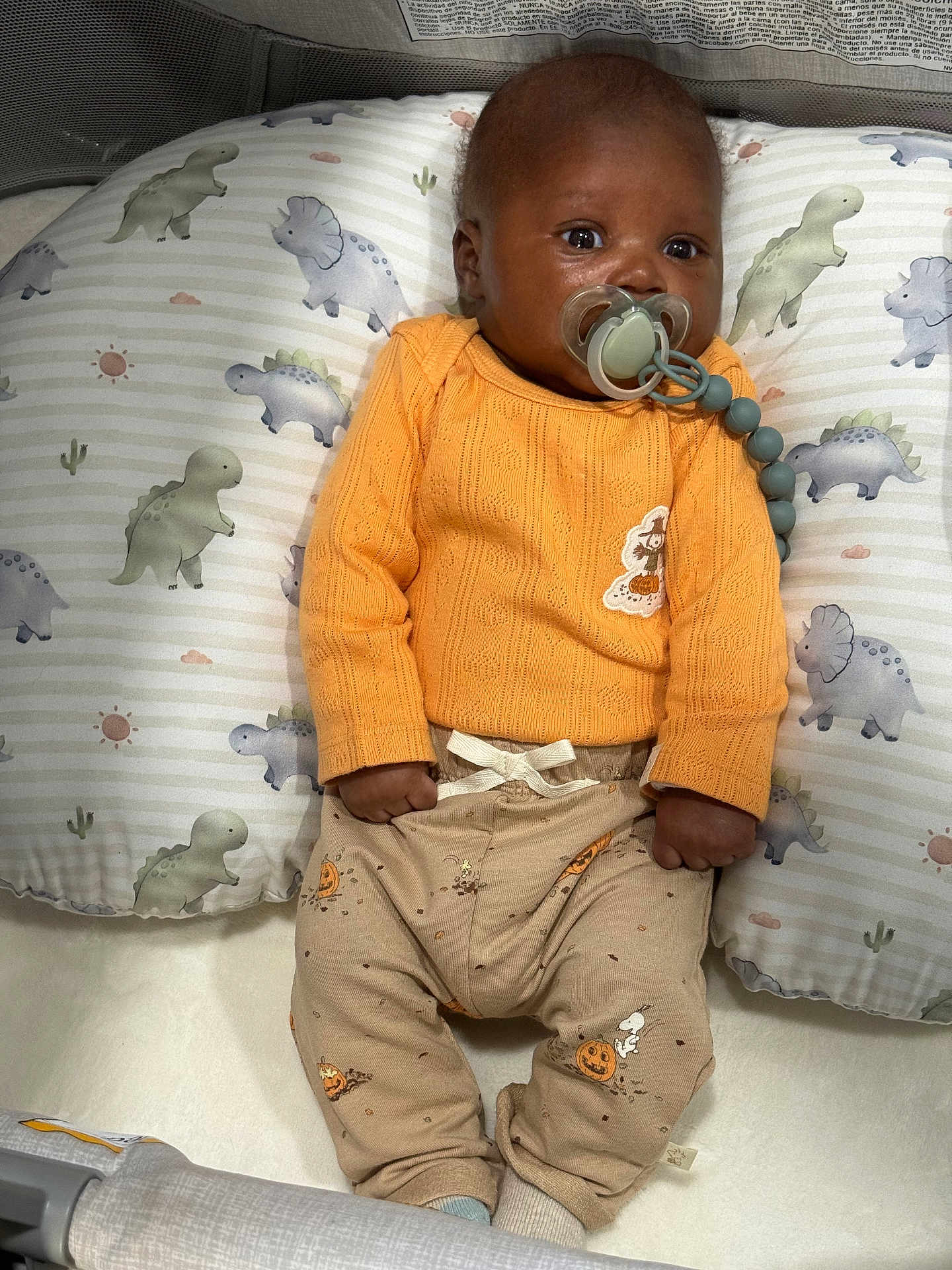 Karter is registered to the contest to win money with this photo: baby, infant, pacifier, orange_sweater, beige_pants, halloween_prints, dinosaur_pillow, cute, indoor, lying_down, child, soft_lighting, closeup, portrait, cozy, baby_clothes, baby_boy, comfort, sleeping_area, playpen