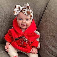 Isabella is registered to the contest to win money with this photo: baby, child, smiling, headband, bow, hoodie, red_clothing, socks, sitting, couch, gray_furniture, indoor, cute, happy, person, infant, portrait, casual, cozy, young